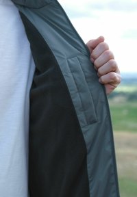 O mână care ține deschis o jachetă închisă la culoare, arătând un buzunar interior neted, purtată peste o cămașă albă, cu un fundal neclar în natură.