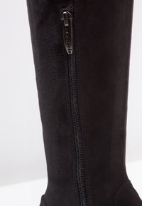 Bota alta de ante negra que cuenta con un cierre lateral y una textura suave. El diseño es elegante con una forma contorneada para un aspecto ajustado.