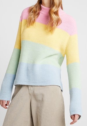 Kobieta ubrana w pastelowy sweter w paski z poziomymi pasami w kolorach różowym, żółtym, miętowym i jasnoniebieskim oraz beżowe spodnie.