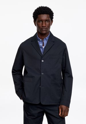 Mand iført en navy blazer over en lyseblå skjorte med knapper, stående med den ene hånd i lommen mod en ensfarvet hvid baggrund.