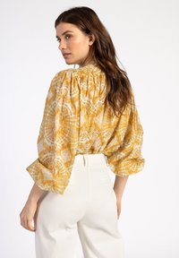 Gelb gemusterte Bluse mit gerafftem Rücken, voluminösen Ärmeln und einem leichten Stoff, kombiniert mit weißen, hochgeschnittenen Hosen.