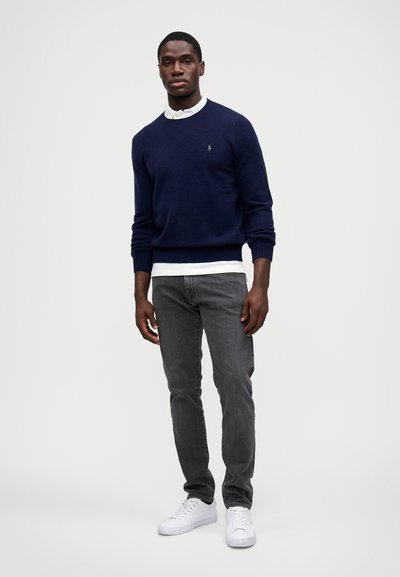 Suéter de malha azul-marinho por cima de uma camisa branca de colarinho, combinado com jeans cinza e ténis brancos. O suéter tem punhos e bainha canelados.