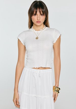 Młoda kobieta ubrana w biały, fakturowany top z krótkim rękawem oraz warstwową spódnicę, ozdobiona złotymi bransoletkami i naszyjnikami, pozuje na jednolitym tle.