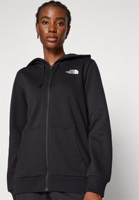 Sudadera con cremallera negra hecha de una tela suave, con un bolsillo frontal y capucha. El logo de The North Face se muestra en blanco en el lado izquierdo del pecho.