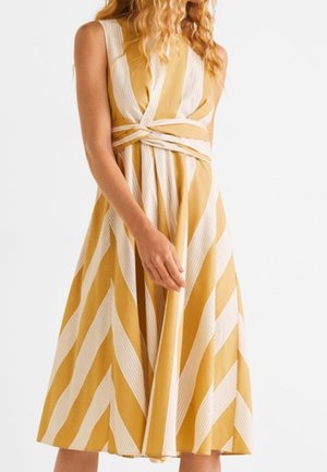 Mouwloze geel-wit gestreepte jurk met diepe V-hals en twistdetail in de taille, gedragen door een staand persoon.