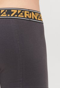 Graue Boxershorts mit einem breiten, schwarz und gelb markierten Bund, der eine glatte Textur aufweist und kontrastierende Nähte entlang der Seiten hat.