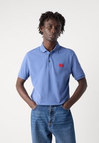 Lys blå polo skjorte med tre knapper, et rødt logo-patch på venstre bryst og korte ermer, kombinert med blå jeans.