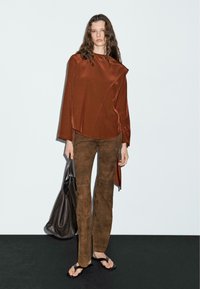 Brun satin top med asymmetrisk design, parret med brune ruskind flared bukser. Modellen holder en mørk lædertaske og har sorte flip-flops på.