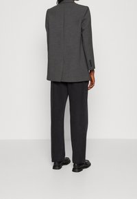 Blazer gris con tejido texturizado, abertura simple en la parte trasera, combinada con pantalones gris oscuro y zapatos negros. Detalle sencillo de botones en las mangas.