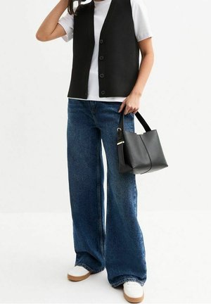 Henkilö, jolla on valkoinen t-paita, musta napillinen liivi, leveälahkeiset siniset farkut, valkoiset kengät, pitää mustaa käsilaukkua remmistä oikeassa kädessä.