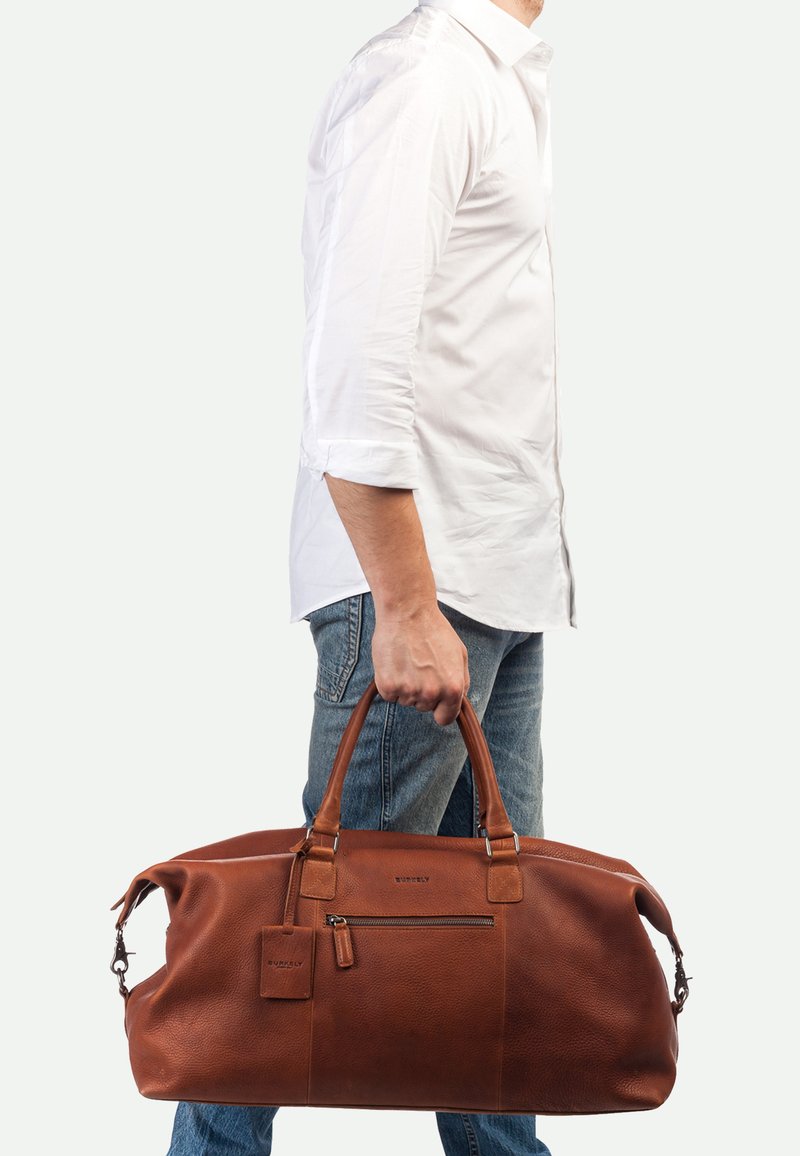 Bruine leren weekendtas met een textuur oppervlak, dubbele handvatten, een ritsvak en een klein label dat eraan is bevestigd.