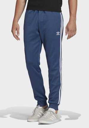 Osoba ubrana w niebieskie spodnie dresowe Adidas z białymi paskami i logo, w połączeniu z beżowymi sneakersami Adidas z białymi paskami.