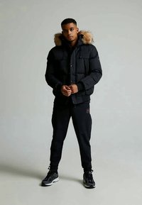 Ung mand står iført en sort puffjakke med pelskrave, sorte joggers og sorte sneakers foran en ensfarvet grå baggrund.