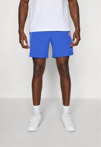 Mann iført blå treningsshorts, hvite sokker, hvite joggesko og en hvit skjorte, stående på en ensfarget lys bakgrunn.