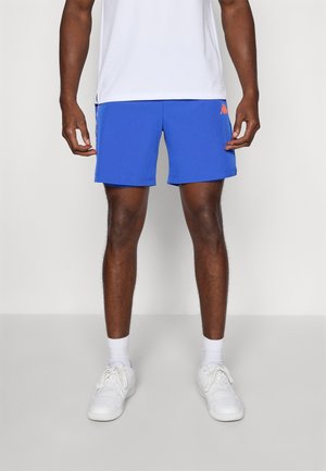 Homme portant un short de sport bleu, des chaussettes blanches mi-mollet, des baskets blanches et un t-shirt blanc, se tenant devant un fond clair.
