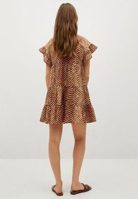 Hnědé šaty s vzorem, s volány na rukávech a vrstvami, které mají cik-cak design na texturované látce. Nosené s hnědými sandály.