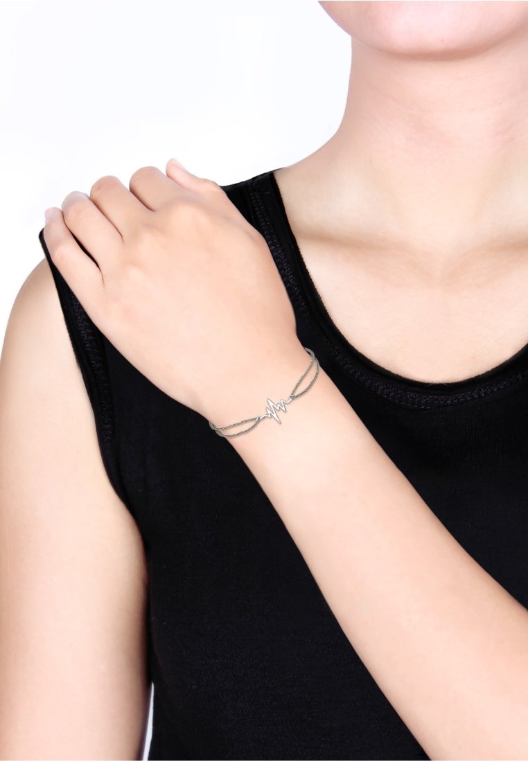 Silberarmband mit einem Herzschlag-Design. Verfügt über verschlungene Stränge mit einer polierten Oberfläche. Wird auf einem nackten Arm zu einem schwarzen Oberteil getragen.