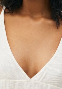 Nahaufnahme einer weiblichen Brust und Nacken, die ein weißes V-Ausschnitt-Oberteil aus geripptem Stoff trägt, mit teilweise sichtbarem lockigem schwarzen Haar.