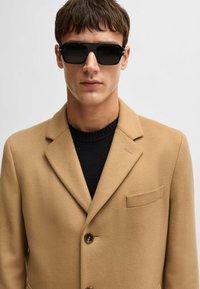 Cappotto in misto lana beige con revers a punta, chiusura a un bottone e tasca sul petto sinistro. Maglione nero a maglia sotto e occhiali da sole neri.