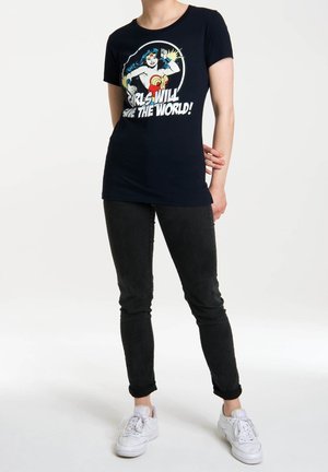 Vrouw draagt zwarte jeans, witte sneakers en een zwart Wonder Woman t-shirt met de tekst "Meisjes zullen de wereld redden!"