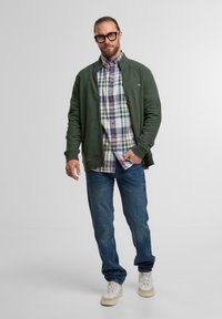 Grüne Reißverschlussjacke über einem karierten Hemd, blaue Jeans und weiße Turnschuhe. Der Stoff wirkt weich und hat eine lockere Passform.