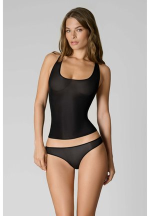 Femme portant un débardeur noir transparent et une culotte noire transparente assortie, debout devant un fond gris uni.