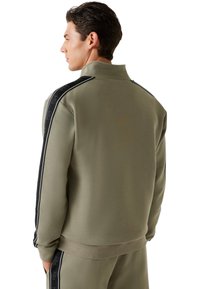 Sudadera verde oliva con cuello alto, tejido texturizado y rayas laterales negras con un patrón. Incluye puños y cintura acanalados.