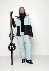 Echipament de schi care include o jachetă impermeabilă bleu deschis și neagră, pantaloni asortați și bocanci de schi închiși la culoare. Modelul ține schiuri negre și mov.