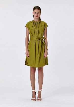 Jeune femme debout portant une robe ceinturée vert olive avec des manches gigot et des talons blancs à bride à la cheville sur un fond blanc uni.