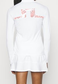 Long-sleeve white shirt with a high collar. Floral pattern in pink on the back. Features a flared skirt hem at the bottom. Soft, smooth texture.