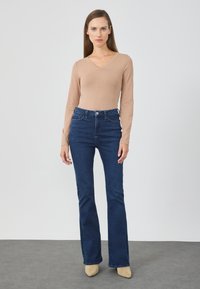 Body beige a maniche lunghe abbinato a jeans a zampa d'elefante a vita alta di colore blu scuro. I jeans hanno tasche frontali e un design aderente. Scarpe neutre.