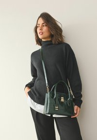 Dark green crocodile-embossed handbag with gold hardware, featuring dual handles and an adjustable shoulder strap. Worn with a black sweater.