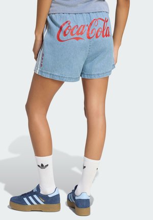 Persona que lleva puestos pantalones cortos vaqueros de color azul claro con el logo rojo de Coca-Cola en la parte trasera, combinados con calcetines blancos de Adidas y zapatillas azules de Adidas.