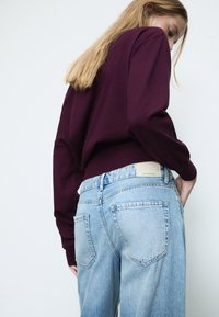 Burgundin neule pitkillä hihoilla, joka on puettu vaaleansinisten korkean vyötärön farkkujen päälle. Farkuissa on klassinen viiden taskun malli ja kevyitä kulutuksia.