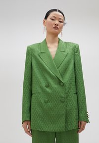 Blazer verde de doble botonadura con un patrón texturizado, solapas con muesca y dos bolsillos frontales. Tejido suave con un ligero brillo.