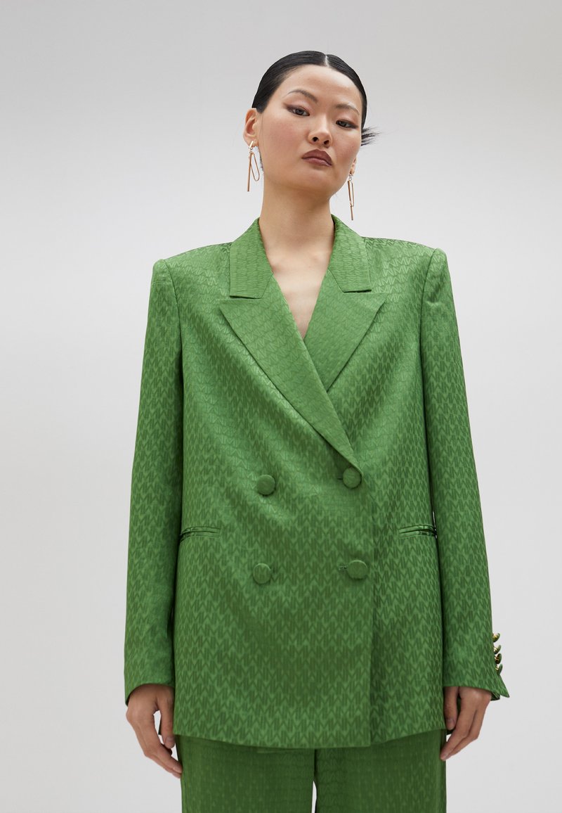 Blazer verde de doble botonadura con un patrón texturizado, solapas con muesca y dos bolsillos frontales. Tejido suave con un ligero brillo.