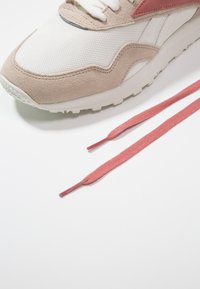 Sneaker con una toma superiore in mesh bianco testurizzato, dettagli in suede beige e una striscia rosa. Presenta lacci piatti rosa posati accanto.