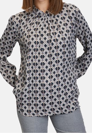 Vrouw draagt een blouse met lange mouwen en een geometrisch diamantpatroon in zwart, wit en grijs, gecombineerd met lichtgrijze broek.