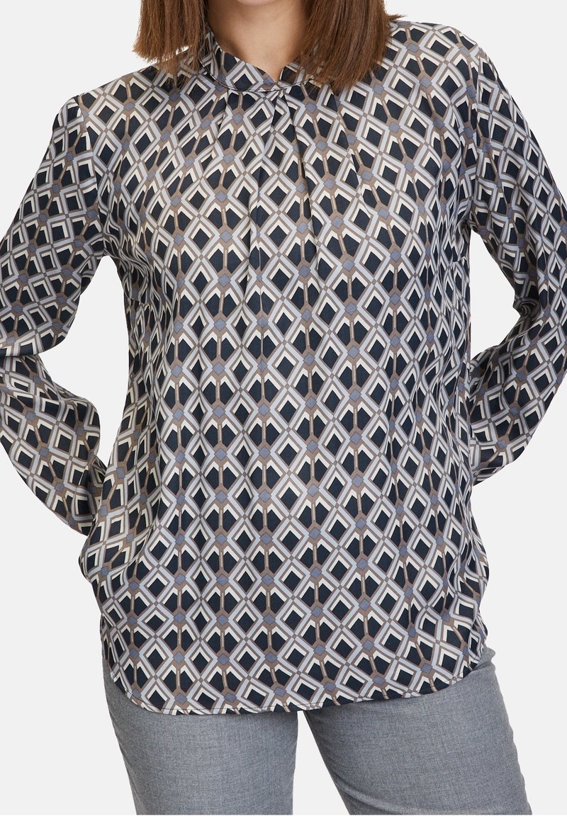 Femme portant un chemisier à manches longues avec un motif géométrique en forme de losange noir, blanc et gris, associé à un pantalon gris clair.