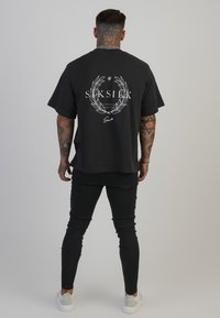Schwarzes Oversized-T-Shirt mit einem weißen Logo und einem Kranz-Graphic auf dem Rücken. Kombiniert mit schwarzen Skinny-Jeans und weißen Sneakers.