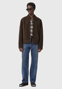 Mand iført en mørkebrun ruskindsjakke, ternet skjorte, blå jeans, sorte støvler og rektangulære sorte solbriller, stående mod en ensfarvet baggrund.