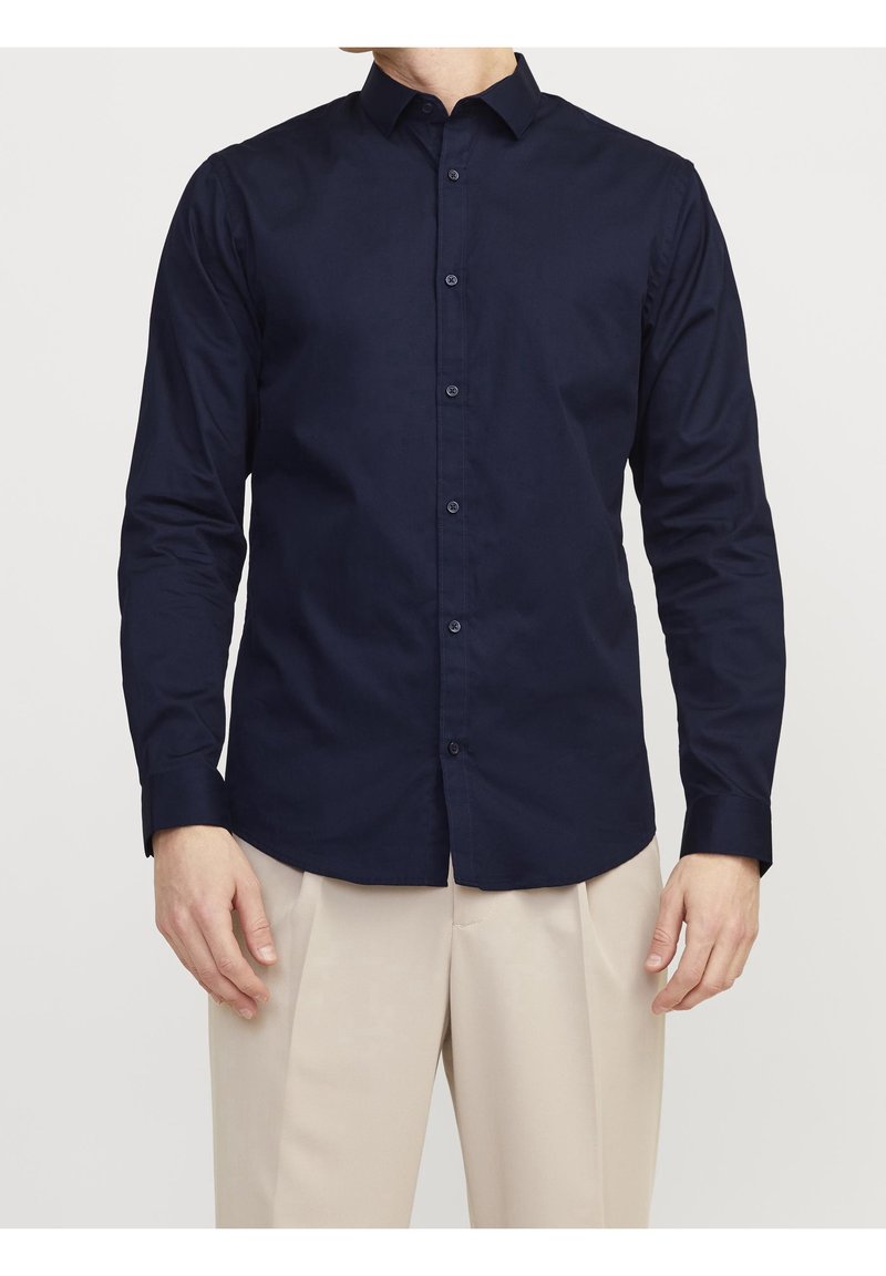 Camisa de manga larga azul marino hecha de algodón, con cuello clásico, cierre de botones en la parte delantera y textura suave. Se lleva con pantalones beige.