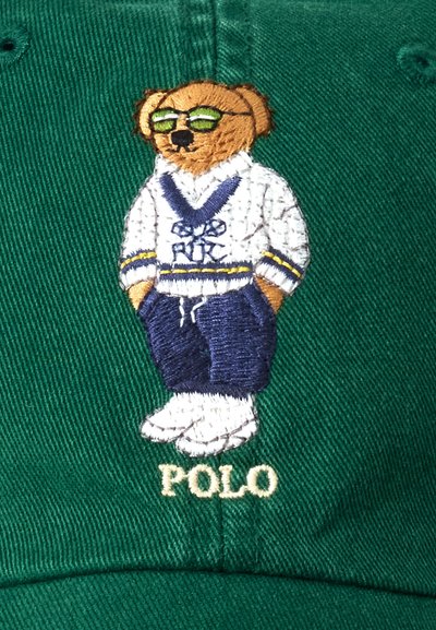 Zöld baseball sapka, amelyen egy hímzett mackó látható, fehér pulcsiban kék részletekkel és sötétkék nadrágban, alatta a "POLO" felirattal.