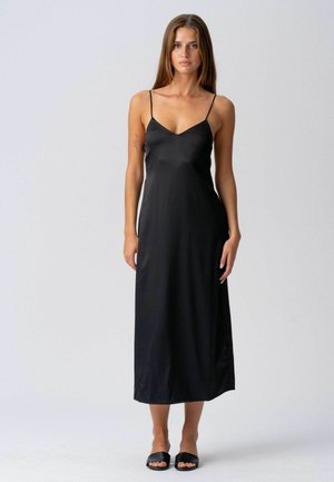 Žena v čiernych midi šatách bez rukávov a v čiernych otvorených šľapkách stojí pred jednoduchým svetlým pozadím.