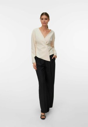 Crème wrap blouse met een diagonale zoom en V-hals, voorzien van lange mouwen en een gouden knoop; gecombineerd met losse zwarte broeken en sandalen.
