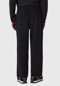 Schwarze Jogginghose aus weichem Stoff mit lockerem Schnitt, elastischem Bund und Seitentaschen. Kombiniert mit schwarzen Turnschuhen mit weißer Sohle.