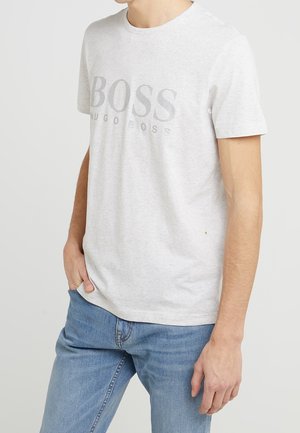 Muž v světle šedém tričku Hugo Boss s logem a modrých džínách, stojící s jednou rukou v kapse proti jednobarevnému pozadí.