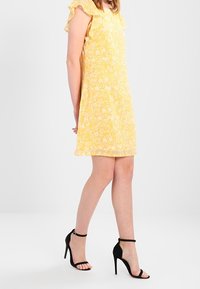 Kvinna i en gul blommig ärmlös klänning och svarta högklackade sandaler med ankelsnörning, står med händerna bakom ryggen.