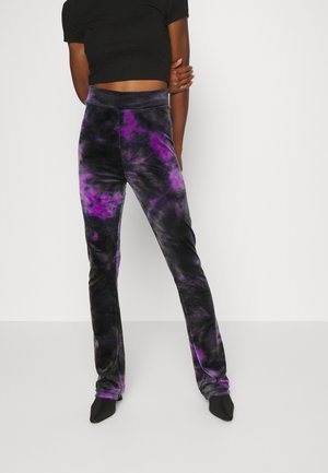 Persoon draagt zwarte en paarse tie-dye broek en een zwart cropped top, staand tegen een effen witte achtergrond.
