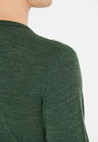Man in een donkergroene, gestructureerde lange mouwen shirt, gezien van achteren met de nek tegen een effen witte achtergrond.